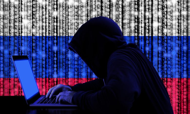 Hacker in a dark hoody sitting in front of a notebook with digital russian flag and binary streams background.