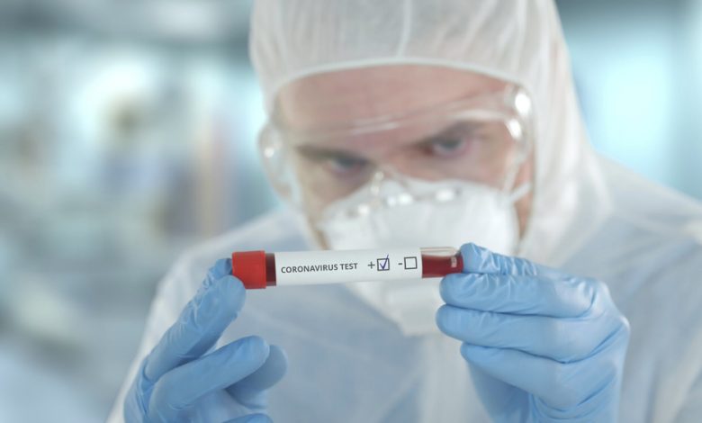 laboratory assistant wearing protection suit examines vial containing a blood sample for COVID tests.