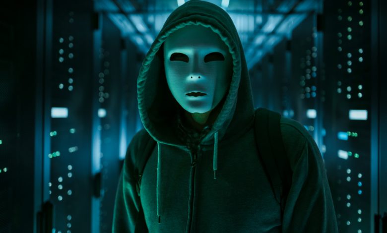 Masked hacker in a hoodie standing in a corporate data center.