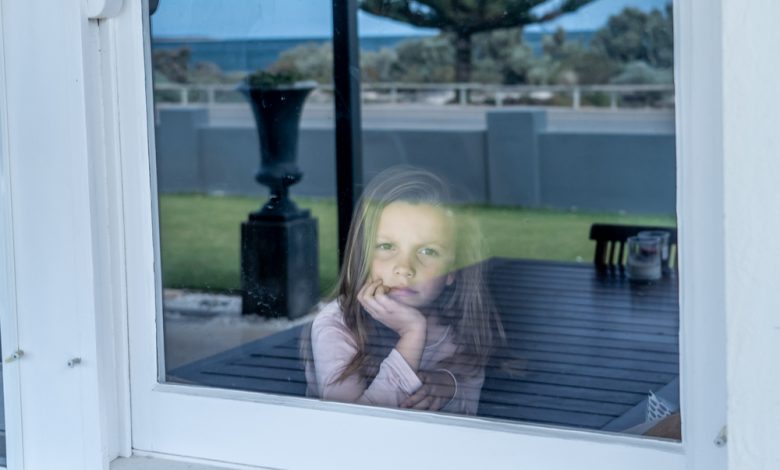 Depressed and lonely little girl looking through the window during COVID-19 quarantine.