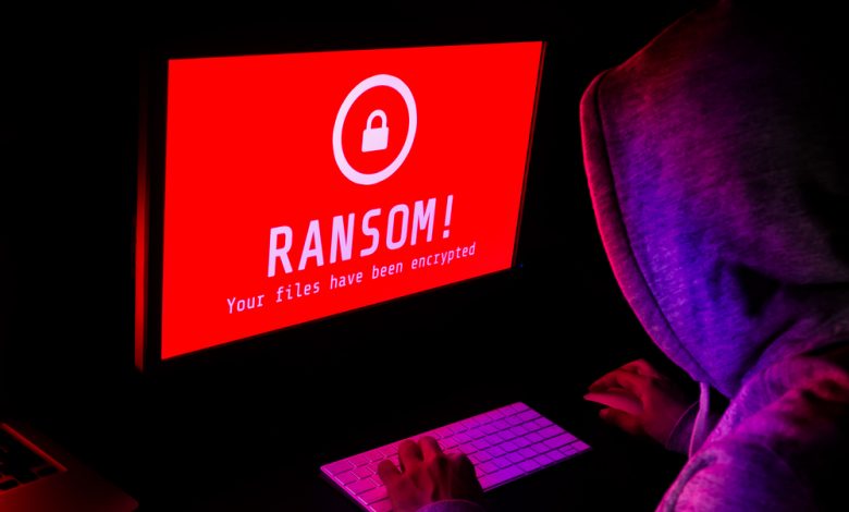Computer screen with ransomware attack alert in red and a hacker man keying on keyboard in a dark room.