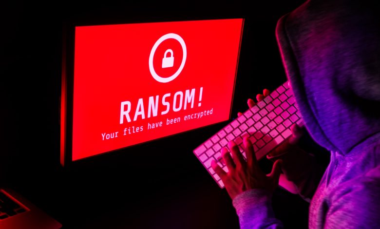 Computer screen with ransomware attack alert in red and a hacker man keying on keyboard in a dark room.