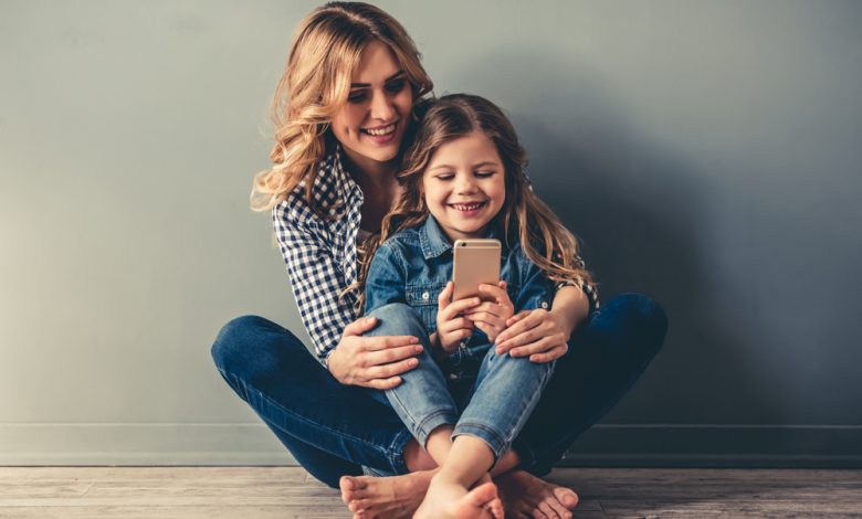 Cute little girl and her beautiful young mom are sitting together on the floor, using a smart phone and smiling, on gray background