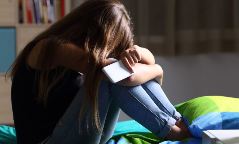 sad teen holding a mobile phone lamenting sitting on the bed in her bedroom