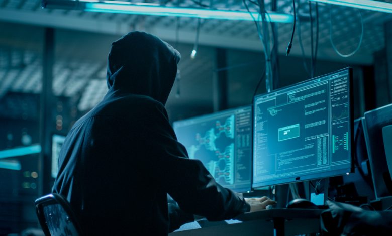 Shot from the Back to Hooded Hacker Breaking into Corporate Data Servers from His Underground Hideout. Place Has Dark Atmosphere, Multiple Displays, Cables Everywhere.