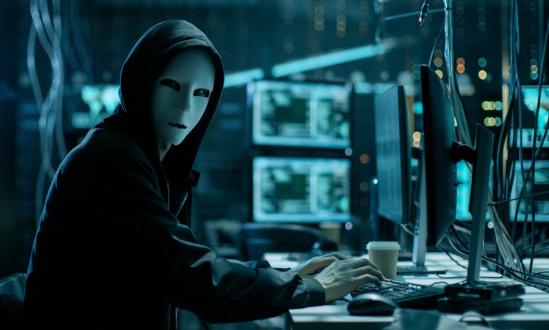 Masked Hacker is Using Computer for Organizing Massive Data Breach Attack on Corporate Servers. They're in Underground Secret Location Surrounded by Displays and Cables.