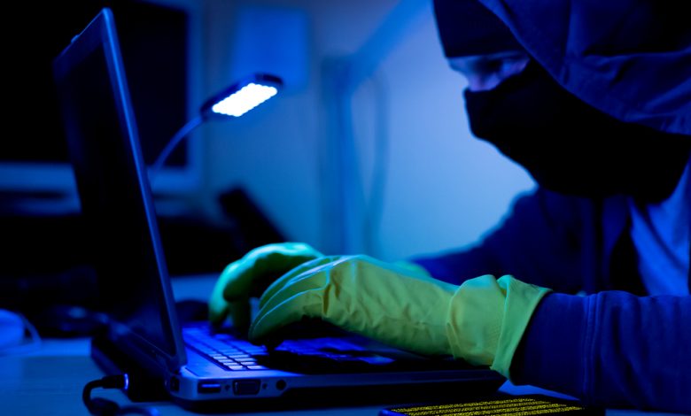 Computer Criminal in balaclava and gloves sitting in the dark room and hacking computer